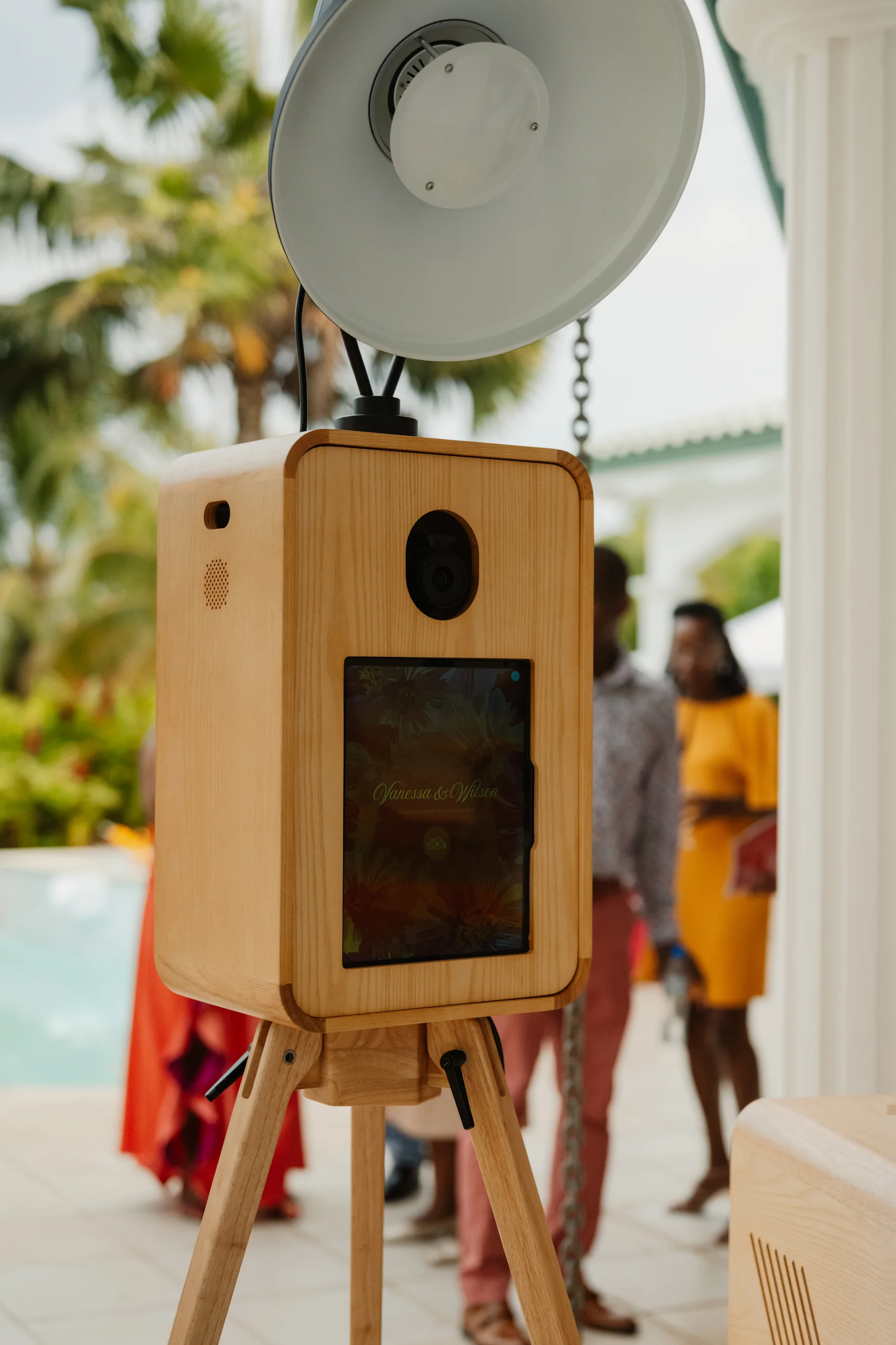 Photobooth BELPOZ sur trépied en bois installé au bord d'une piscine, invités en arrière-plan